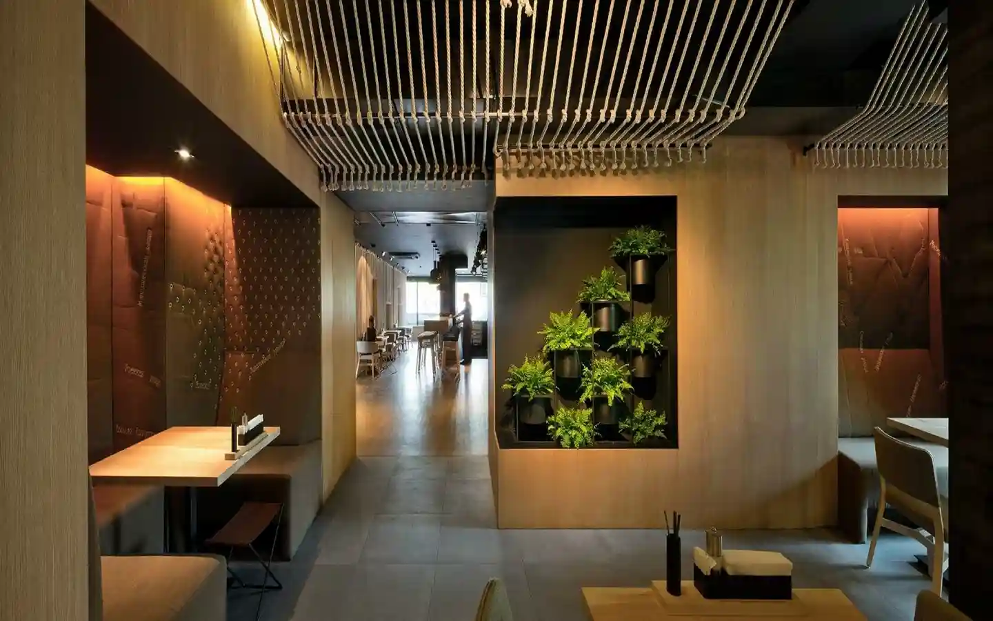 Modern restaurant interior featuring a vertical indoor plant display, rope-textured ceiling installation, and cozy recessed booth seating