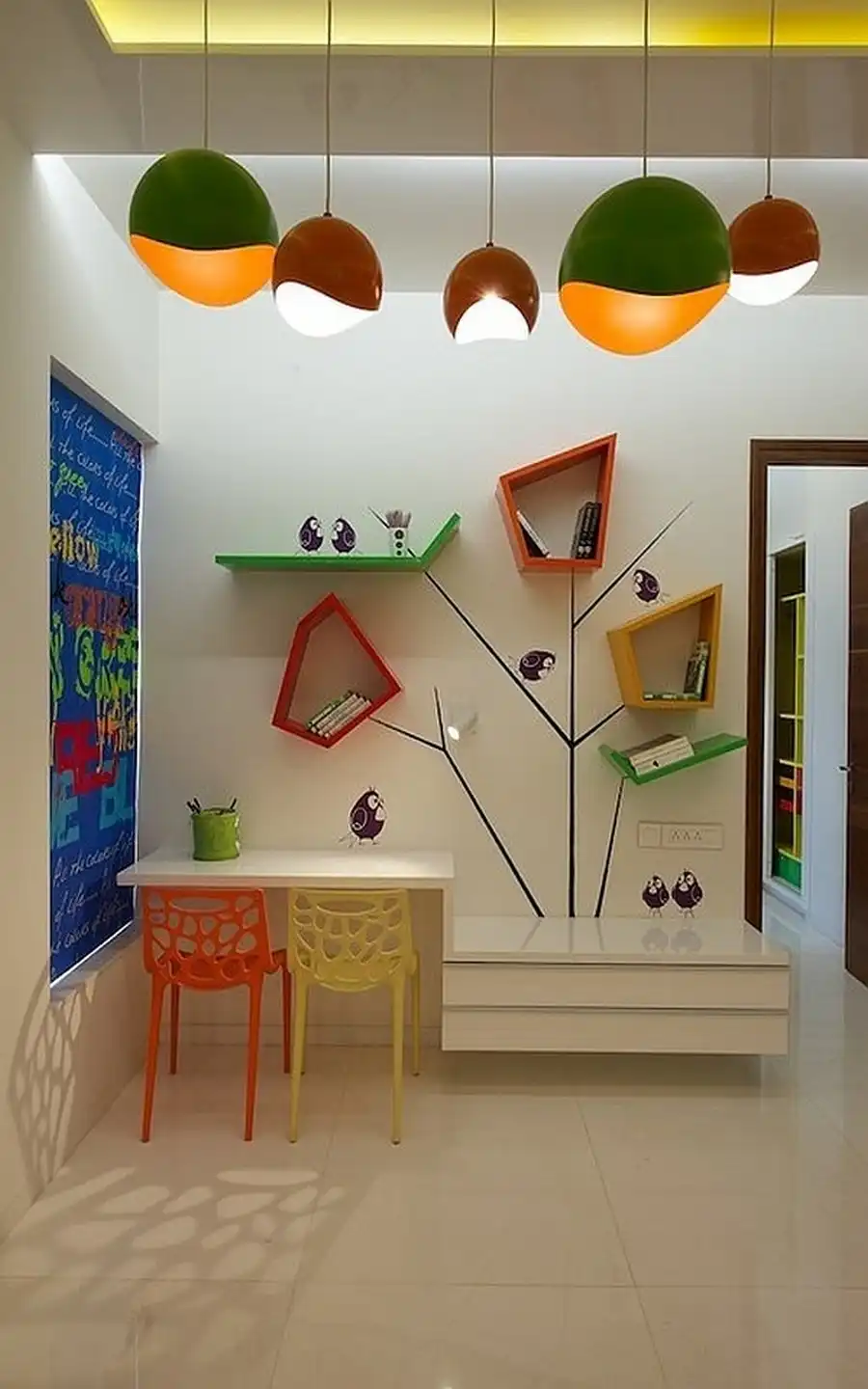 Colorful kids' study room featuring a tree-shaped wall bookshelf with geometric cubbies, modern orange and yellow honeycomb chairs, and vibrant spherical pendant lights