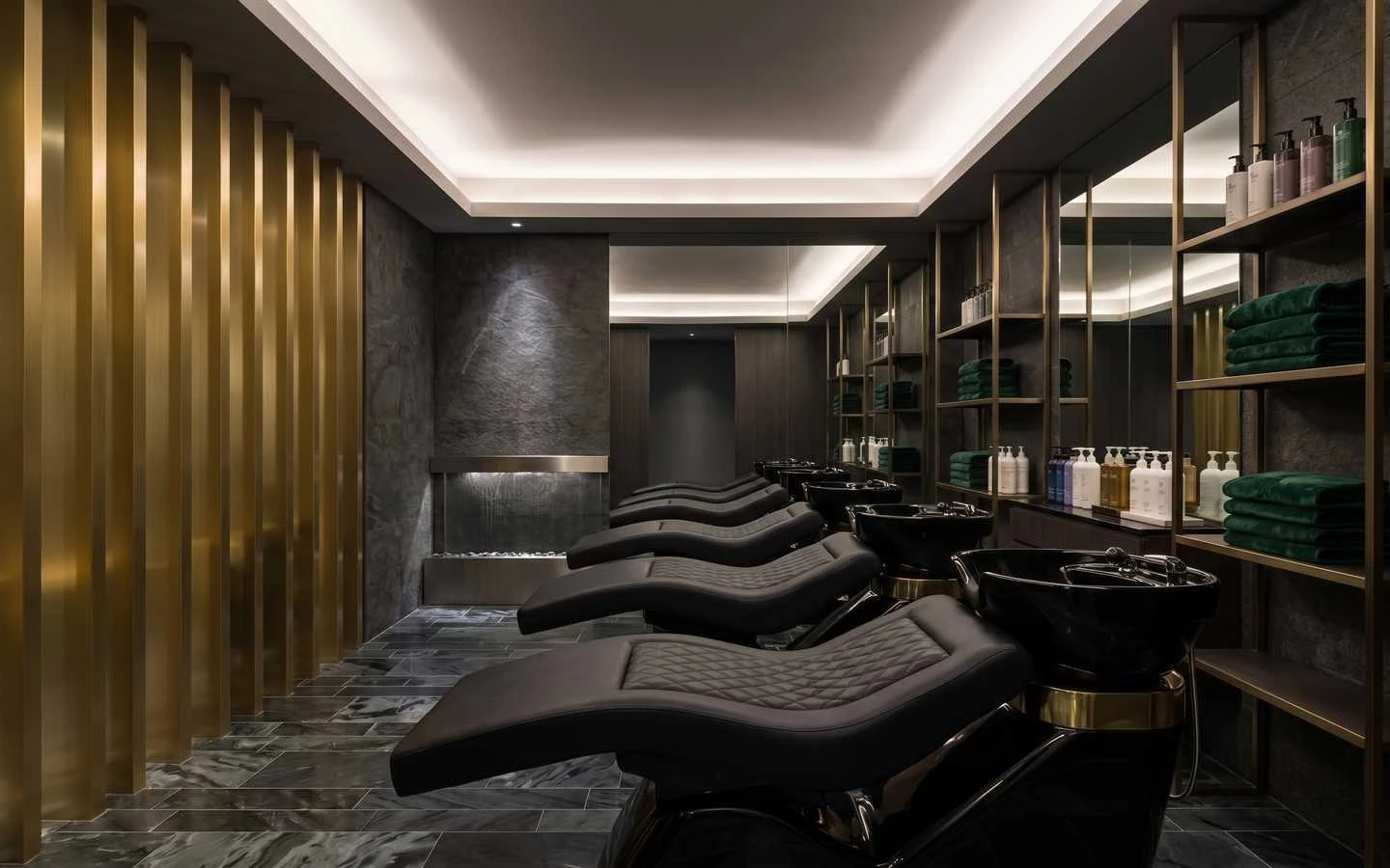 Luxury salon shampoo area with black quilted reclining chairs, gold vertical wall slats, dark stone walls, and ambient cove lighting