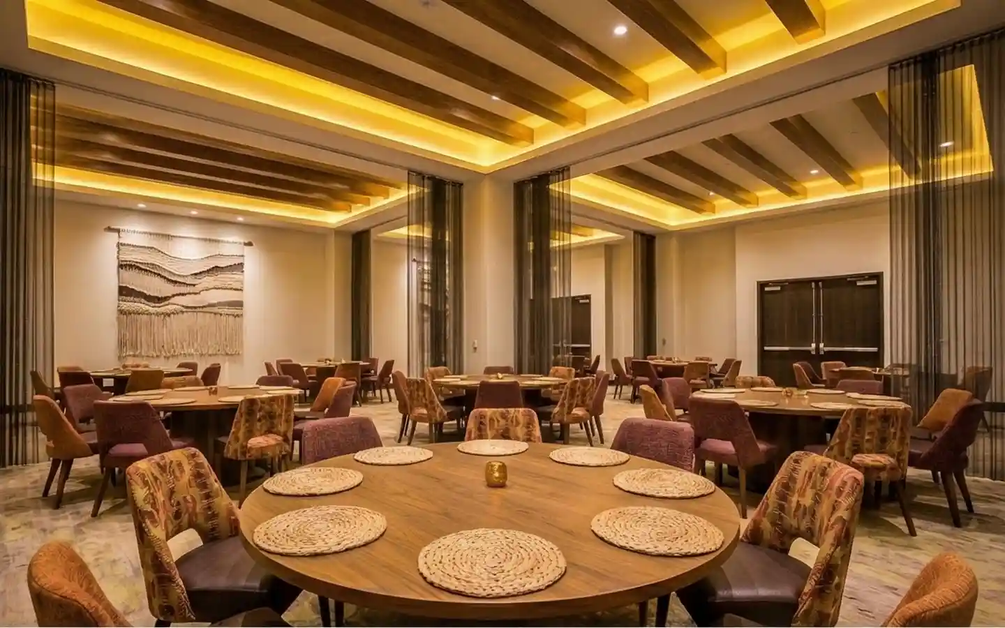 Elegant hotel banquet hall featuring a wood beam ceiling with warm cove lighting, round dining tables, and patterned designer chairs