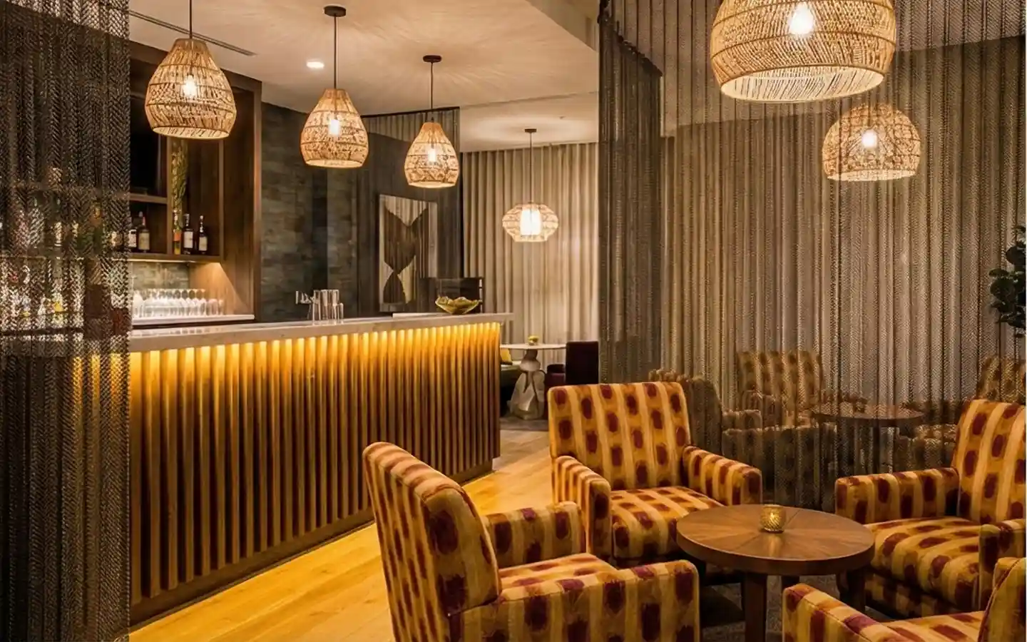 Cozy hotel bar lounge featuring a warm backlit counter, rattan pendant lights, patterned armchairs, and mesh room dividers