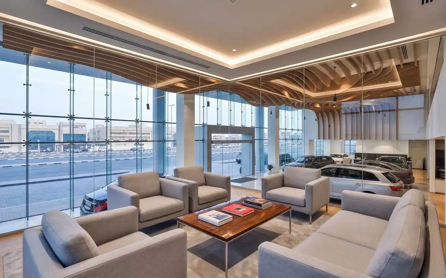 Modern commercial interior design of a car showroom lounge featuring gray minimalist armchairs, a wood coffee table, and a view of high-end vehicles through glass partitions