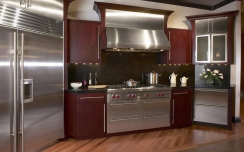 Luxury kitchen interior featuring dark cherry wood cabinetry, professional-grade stainless steel appliances including a double oven and large refrigerator, a black backsplash, and warm under-cabinet lighting