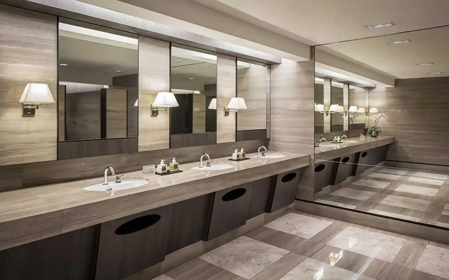 Luxury commercial restroom interior design featuring a long stone vanity with multiple sinks, individual mirrors with elegant wall sconces, and a patterned marble tile floor