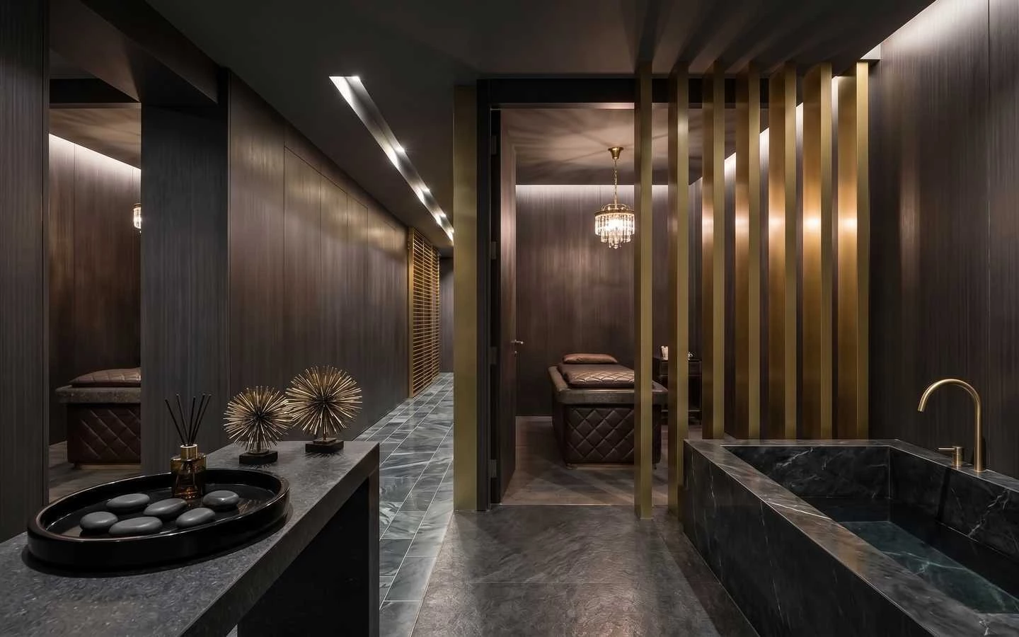 Luxury dark-themed spa interior featuring a black marble soaking tub, gold slat room dividers, quilted massage tables, and elegant crystal chandelier lighting