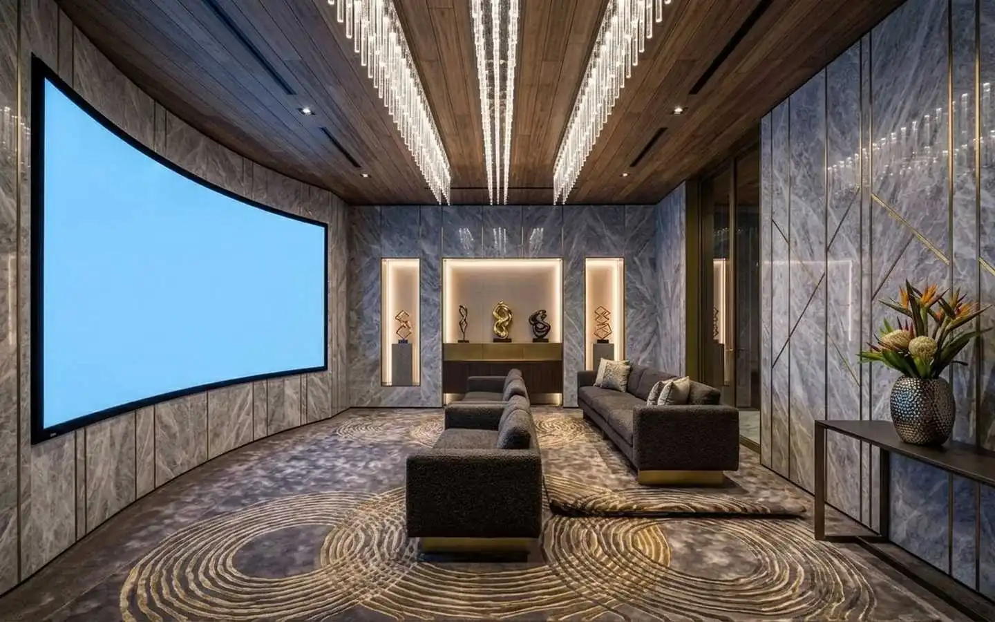 Modern luxury home theater featuring a large curved cinema screen, marble-cladded walls, a wood-slat ceiling with crystal chandeliers, and plush grey seating