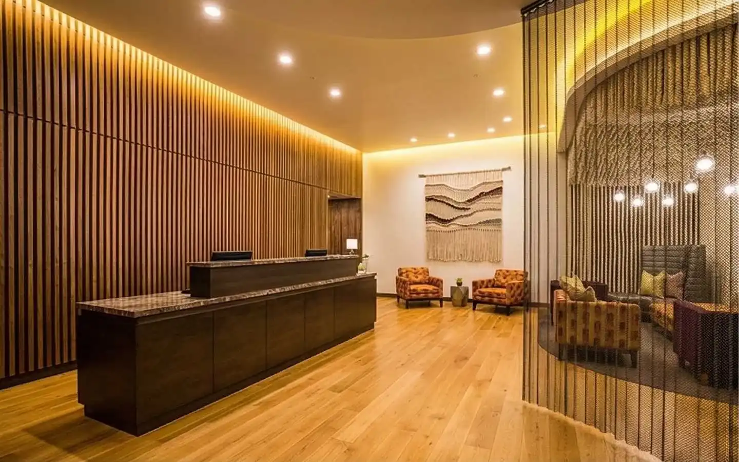 Luxury hotel reception lobby featuring a dark wood front desk, vertical wood slat wall, warm ambient lighting, and a cozy lounge area with patterned armchairs