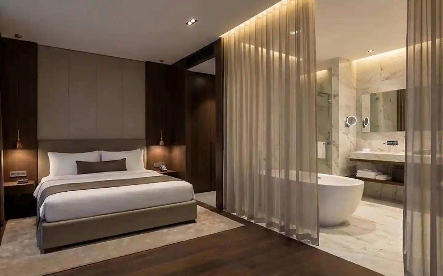 Luxury hotel bedroom and open marble bathroom separated by elegant floor-to-ceiling sheer curtains, featuring a freestanding tub and warm cove lighting