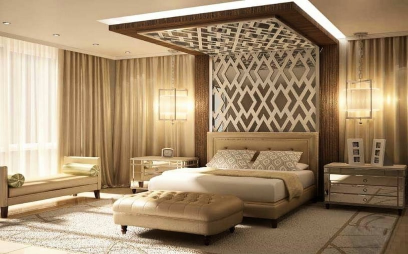 Luxury master bedroom featuring a decorative lattice wood ceiling and wall panel, mirrored nightstands, a tufted ottoman bench, and elegant cream-colored drapes