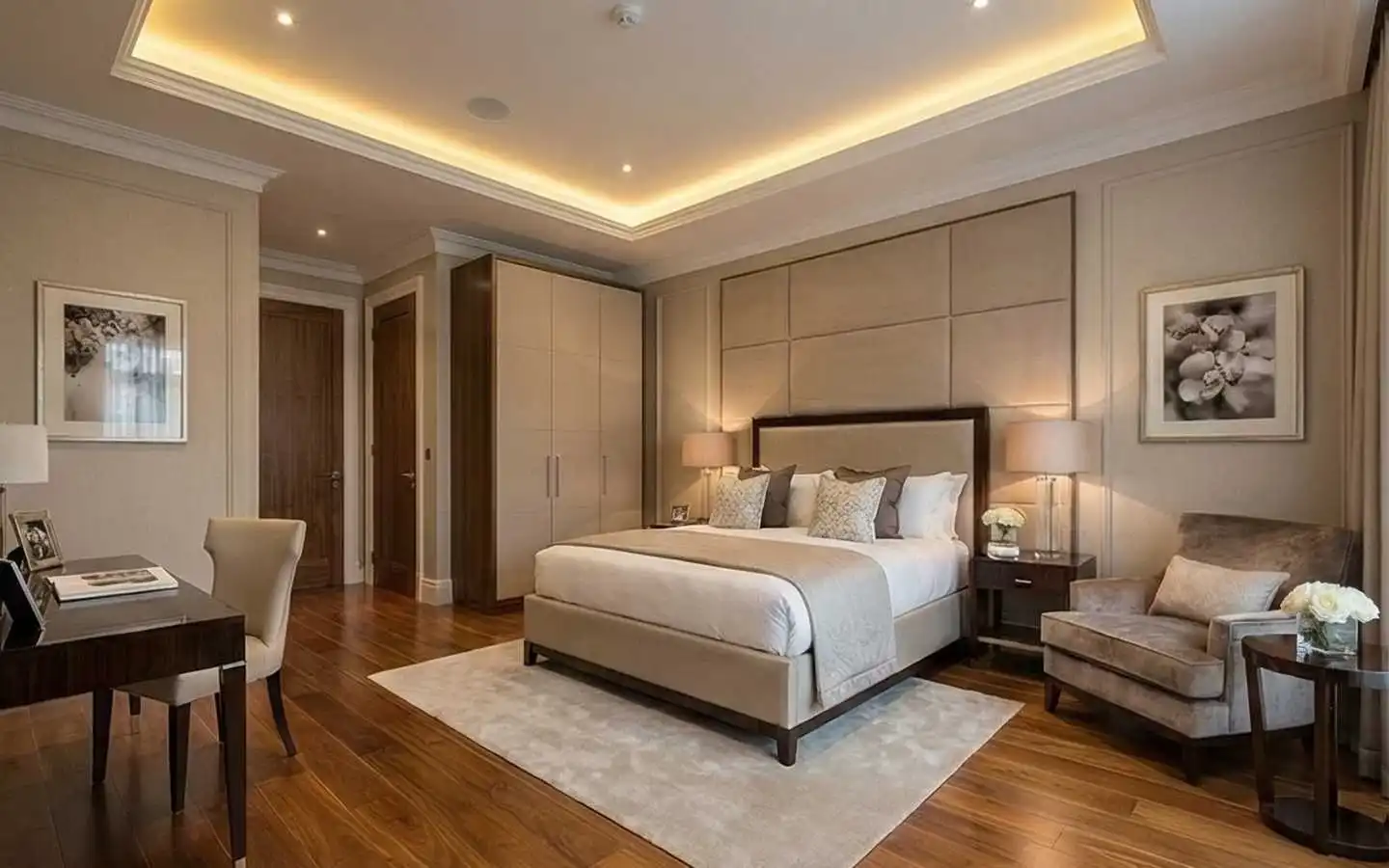 Elegant master bedroom featuring a large upholstered wall panel headboard, hardwood floors, warm cove ceiling lighting, and a dark wood vanity desk