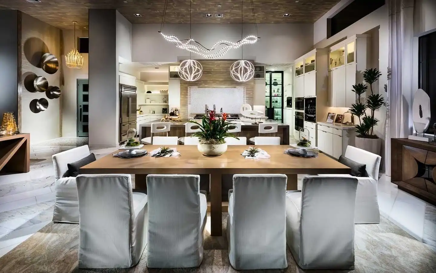 Luxury open-concept kitchen and dining area featuring a large wooden table with white slipcovered chairs, avant-garde LED pendant lighting, and a high-end chef's kitchen