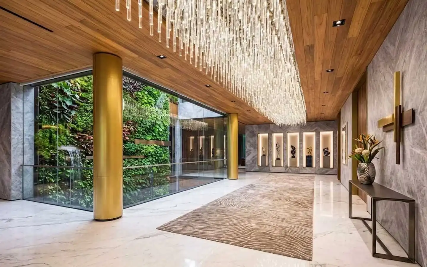 Opulent villa hallway featuring a long crystal fringe chandelier, gold structural pillars, marble walls with art niches, and a glass-enclosed vertical garden with a waterfall