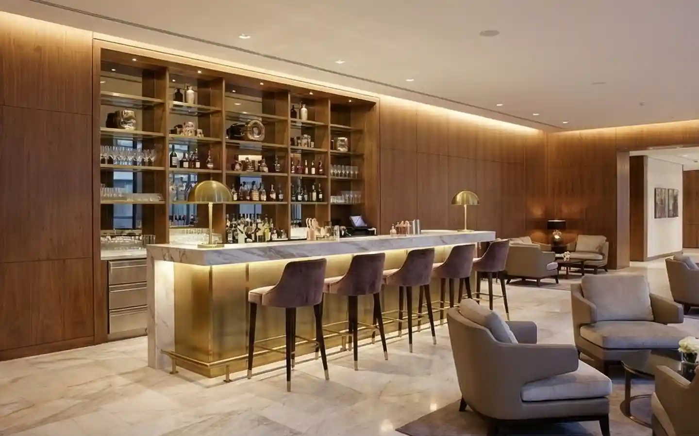 Luxury hotel bar interior featuring a white marble countertop with integrated gold lighting, velvet upholstered high chairs, and floor-to-ceiling wood wall cladding
