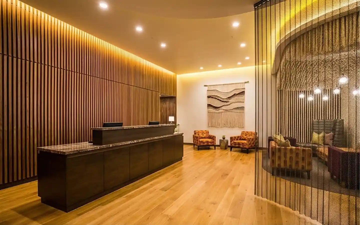 Hotel reception desk with a vertical wood slat feature wall, hardwood floors, and a decorative metal mesh screen separating the lounge seating area
