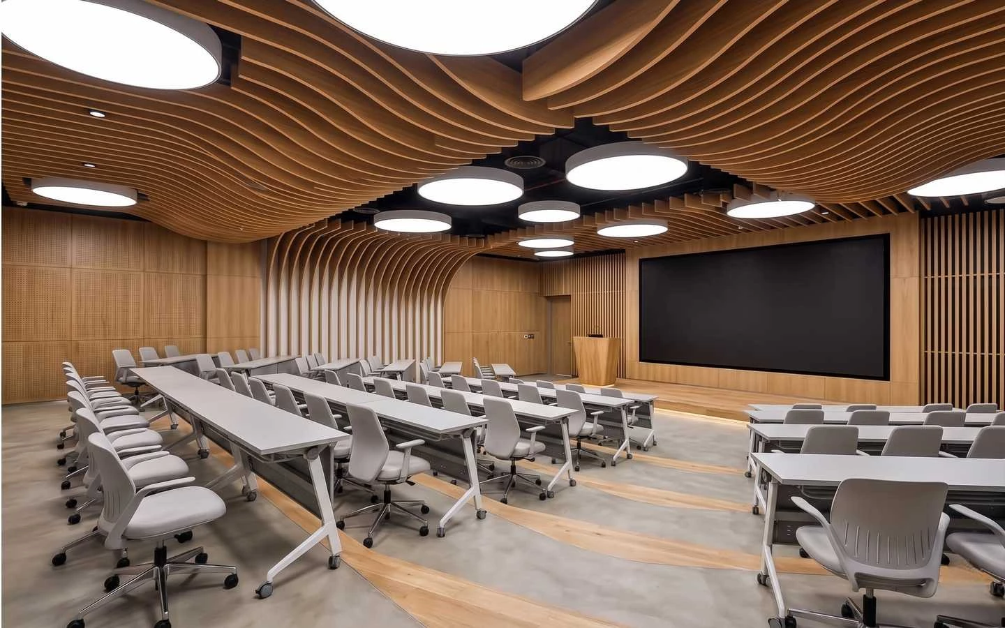 Modern office training room with a wave-patterned wooden baffle ceiling, tiered seating with white desks, and a large presentation screen