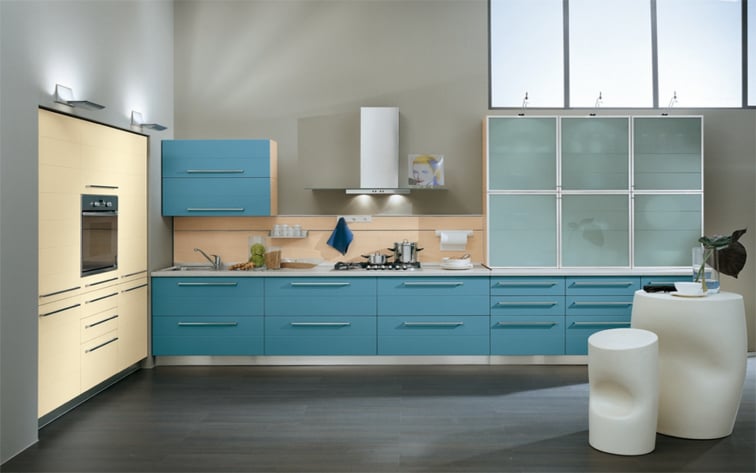 Contemporary modular kitchen featuring teal blue base cabinets, cream-colored pantry storage, and frosted glass wall units with a minimalist white breakfast table