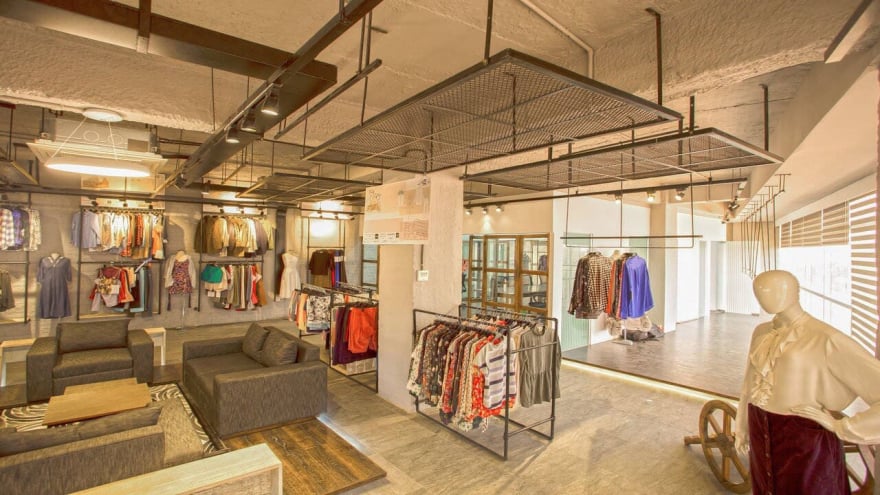 Modern boutique clothing store interior with wall-mounted and freestanding apparel racks, mannequins, seating area with couches, and industrial-style ceiling lighting