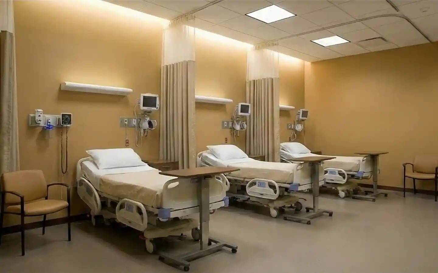 Professional clinic recovery room with adjustable medical beds, integrated wall-mounted monitors, privacy curtains, and warm neutral wall tones