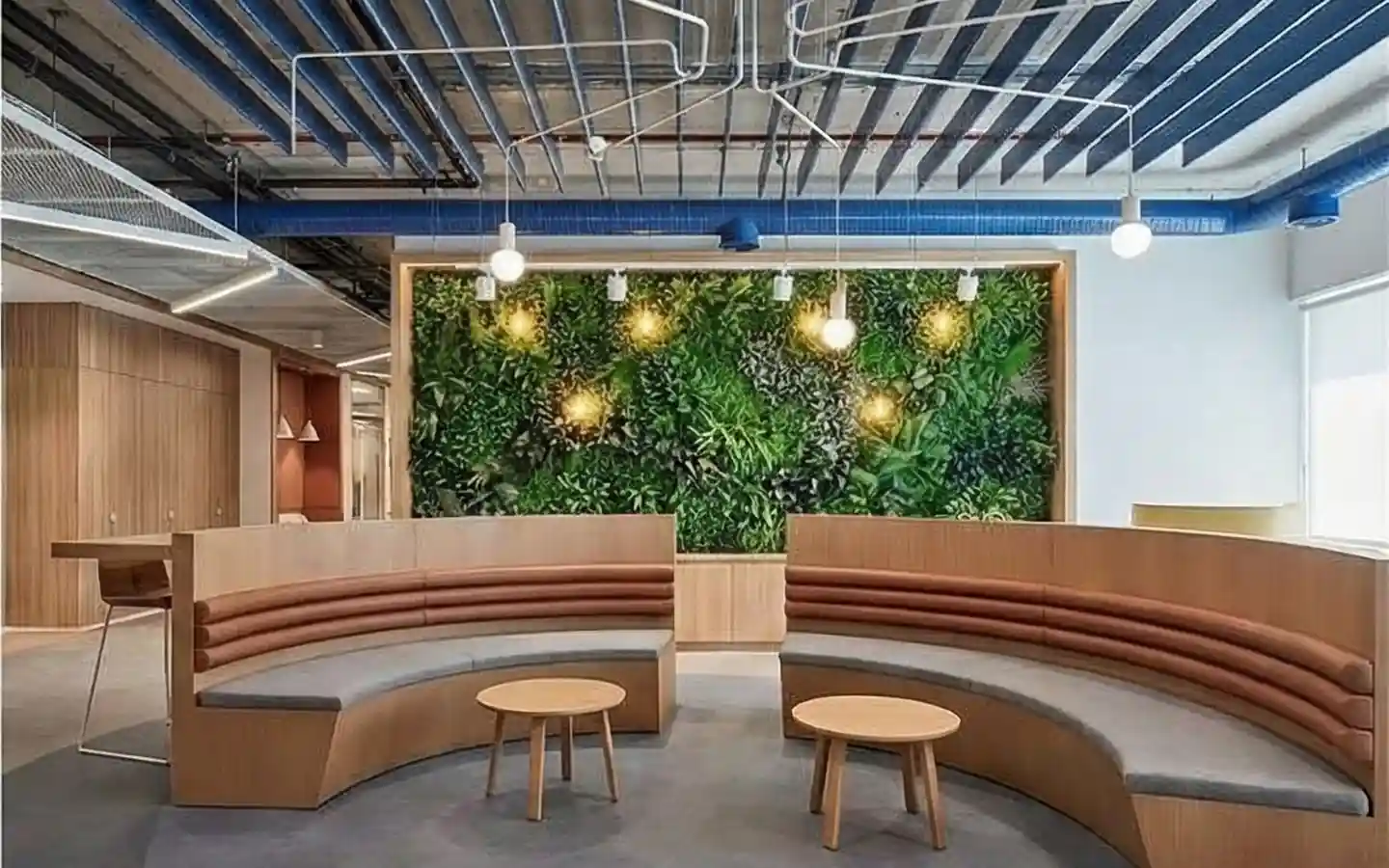Modern clinic waiting space featuring a lush vertical plant wall, curved wooden banquet seating, and an industrial open-grid ceiling with blue piping