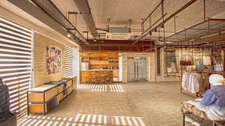 Spacious modern clothing showroom interior with mannequins, apparel racks, wooden storage units, natural light through striped blinds, and a small coffee/bar area