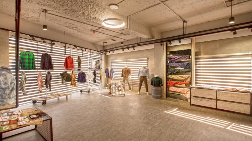 Modern clothing store interior with apparel displayed on wall-mounted racks, mannequins showcasing outfits, folded clothes on shelves, and natural light streaming through striped blinds