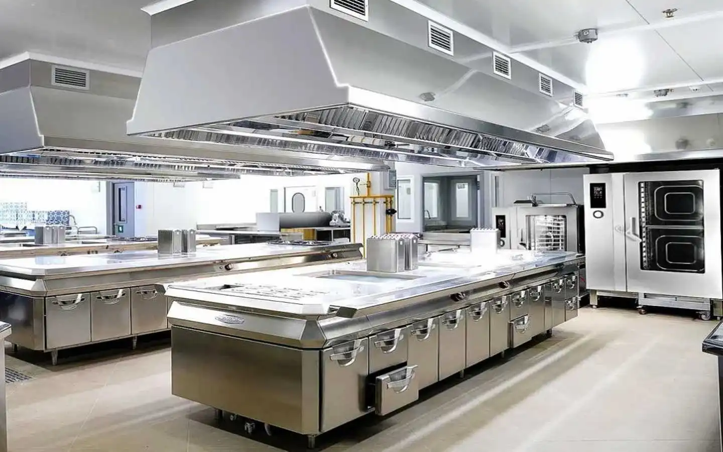 Modern commercial kitchen interior design featuring large stainless steel prep islands, heavy-duty ceiling ventilation hoods, and industrial-grade cooking equipment
