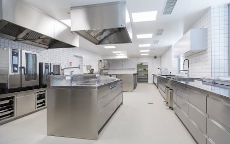 Clean and modern commercial kitchen interior featuring stainless steel prep stations, industrial ovens, large ventilation hoods, and white tiled walls