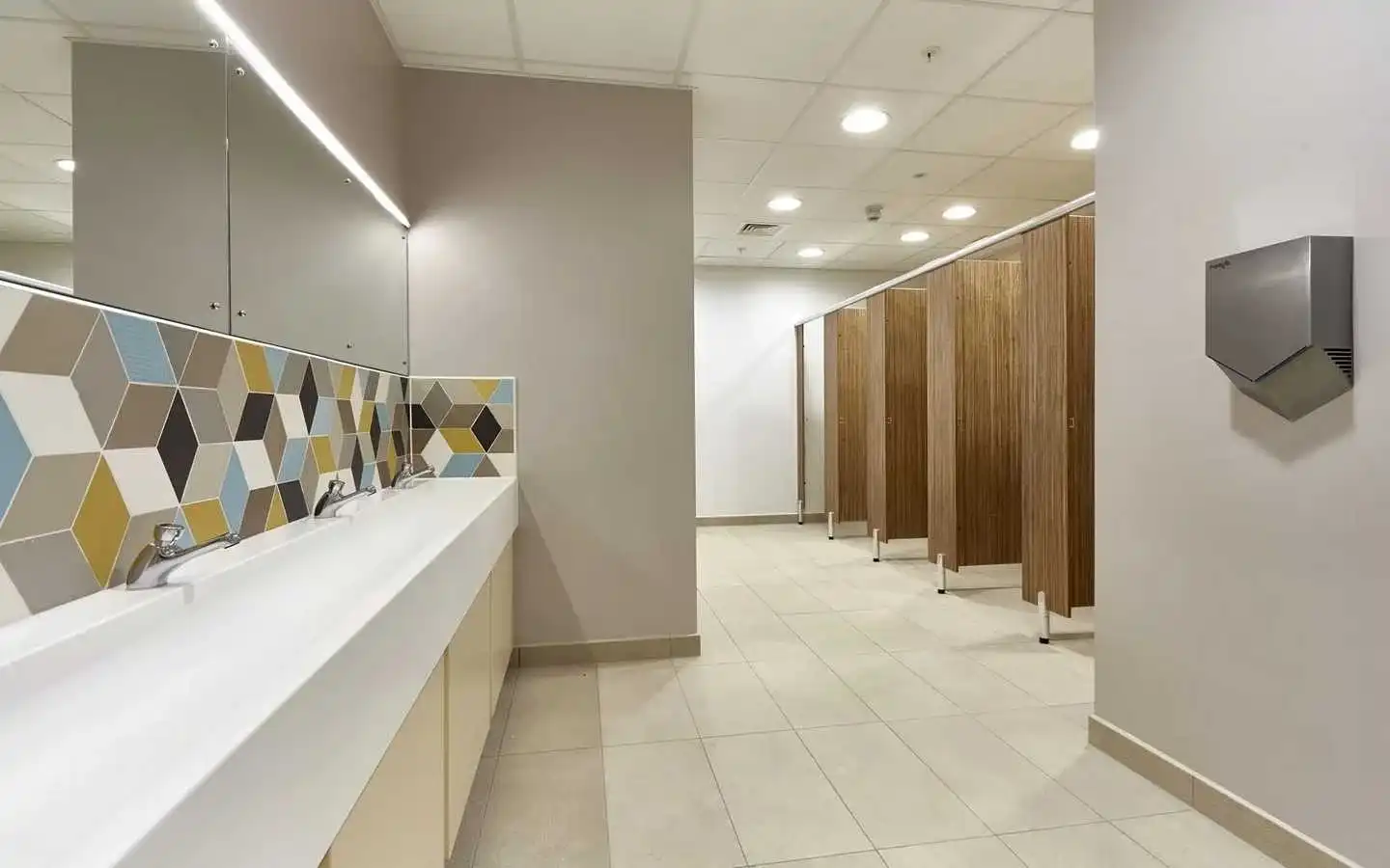 Modern commercial washroom interior featuring a white trough sink, geometric 3D-effect backsplash tiles, large mirrors with LED strip lighting, and wood-finish toilet cubicles
