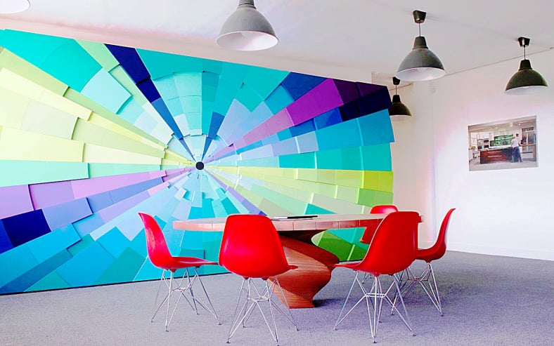 Vibrant modern conference room featuring a large 3D abstract geometric wall mural in blue and green hues, red mid-century modern chairs, and a sculptural wooden table