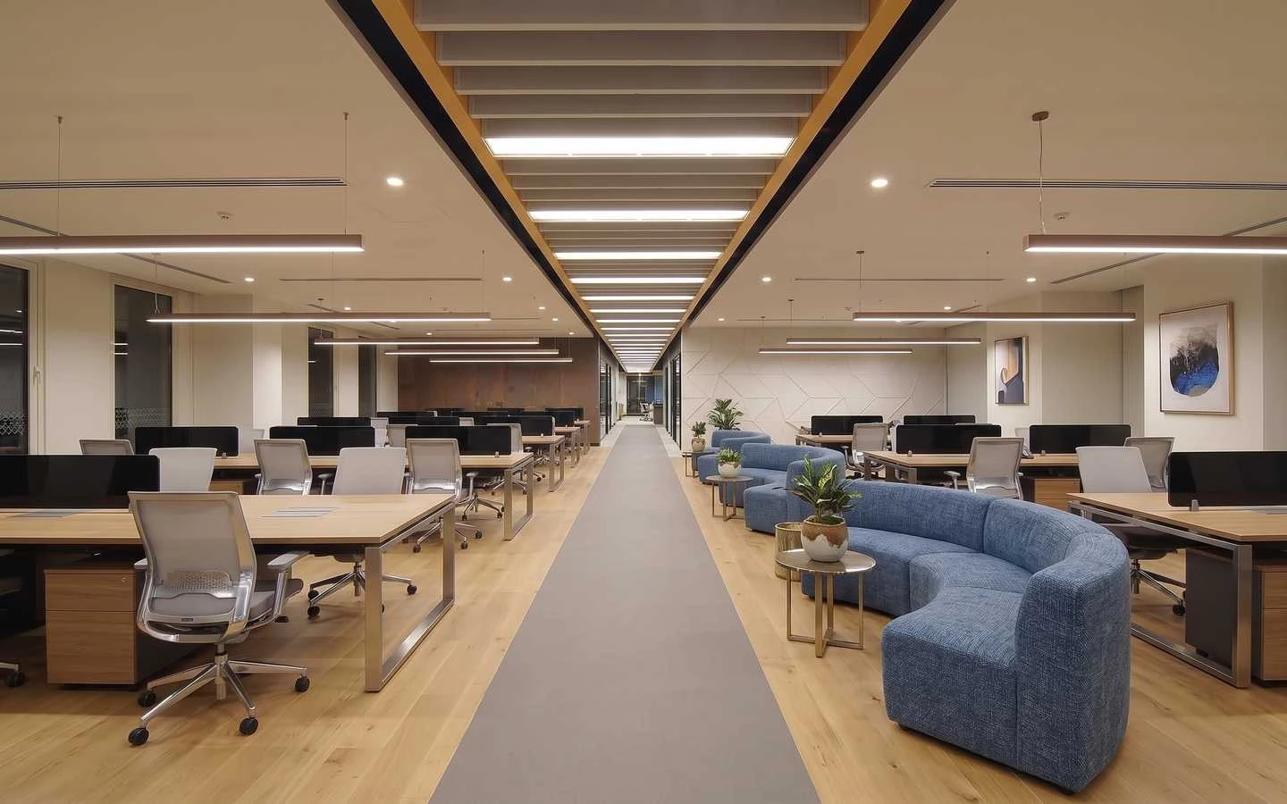 Spacious modern corporate office interior with long rows of wooden desks, ergonomic chairs, curved blue lounge sofas, and linear pendant lighting