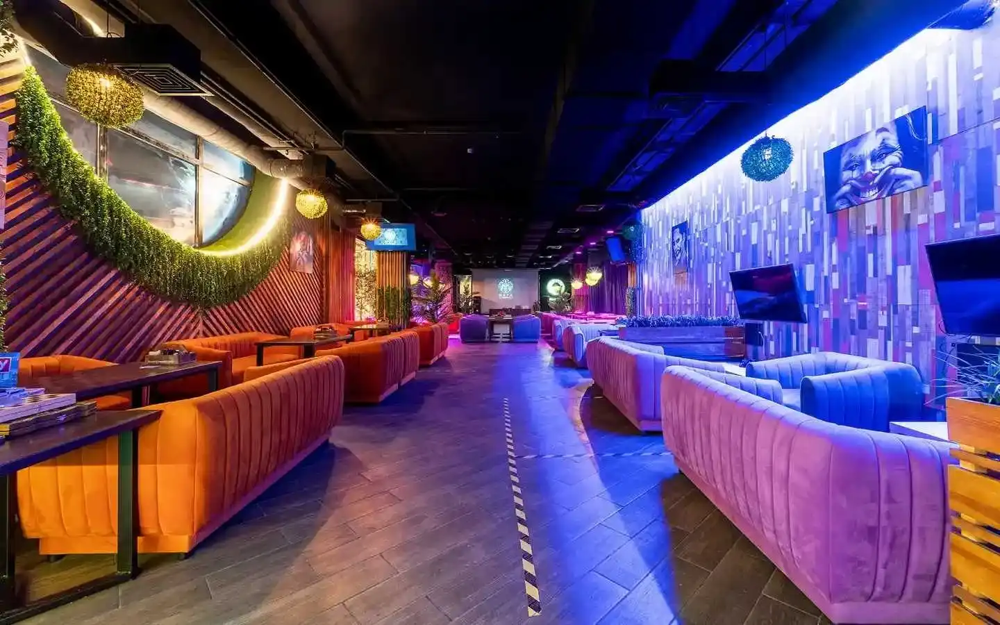 Modern entertainment lounge interior featuring vibrant orange and purple velvet sofas, neon accent lighting, decorative greenery, and patterned wall art