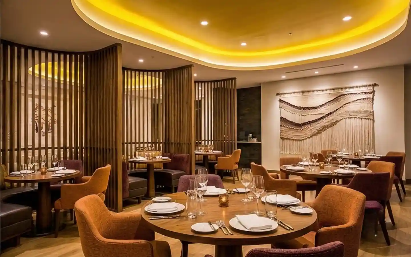 Modern hotel restaurant interior featuring curved wood slat partitions for private booth seating, warm cove lighting, and round wooden dining tables