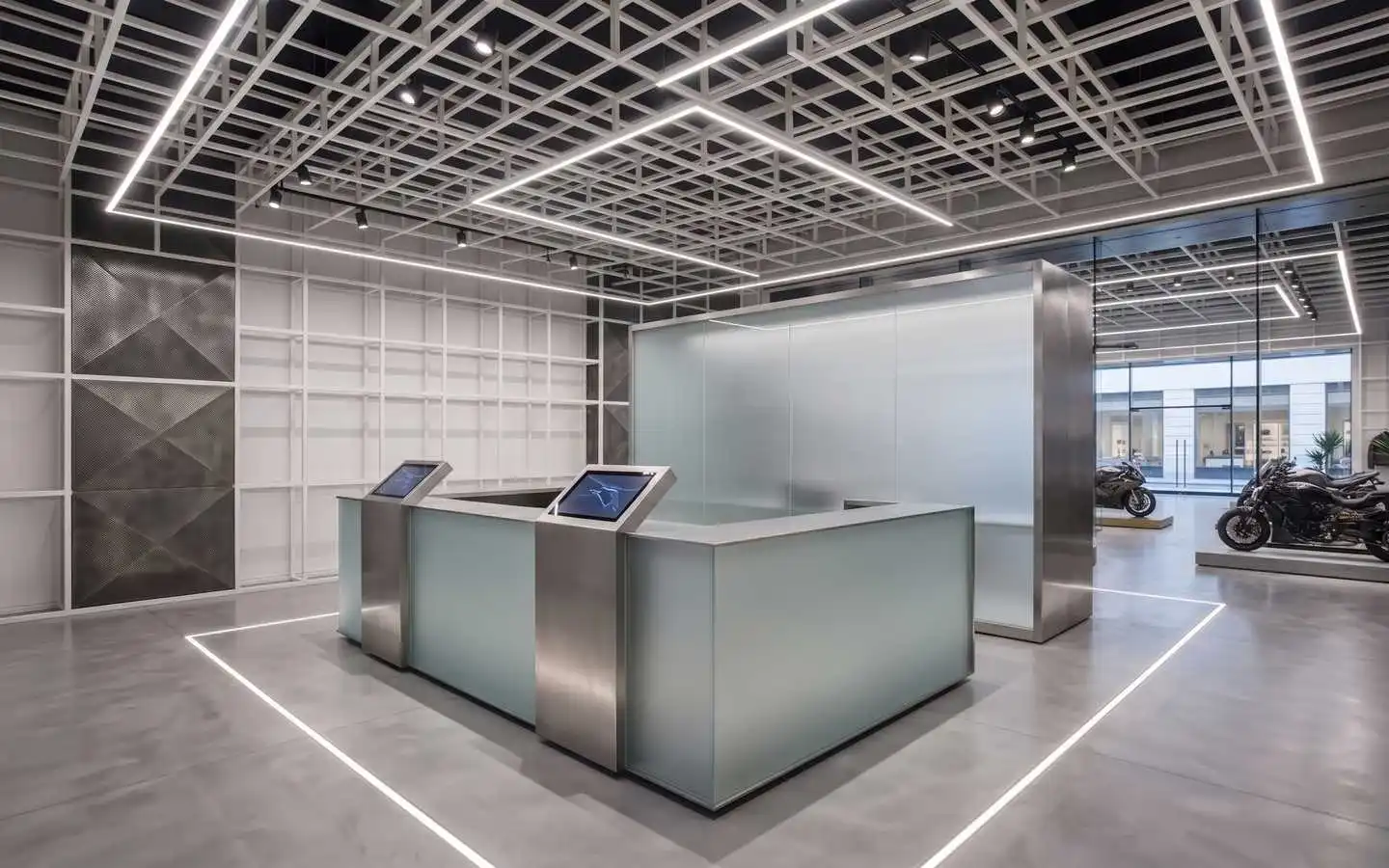 Modern industrial showroom interior featuring a frosted glass reception desk with digital displays, white grid ceiling with linear LED lighting, and polished concrete floors