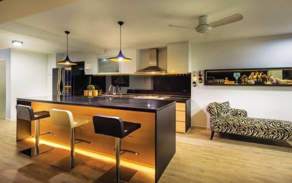 Modern open-concept kitchen interior featuring a large island with black countertops and warm under-lighting, three adjustable bar stools, black pendant lights, and a distinct zebra-print lounge chair in the adjoining space