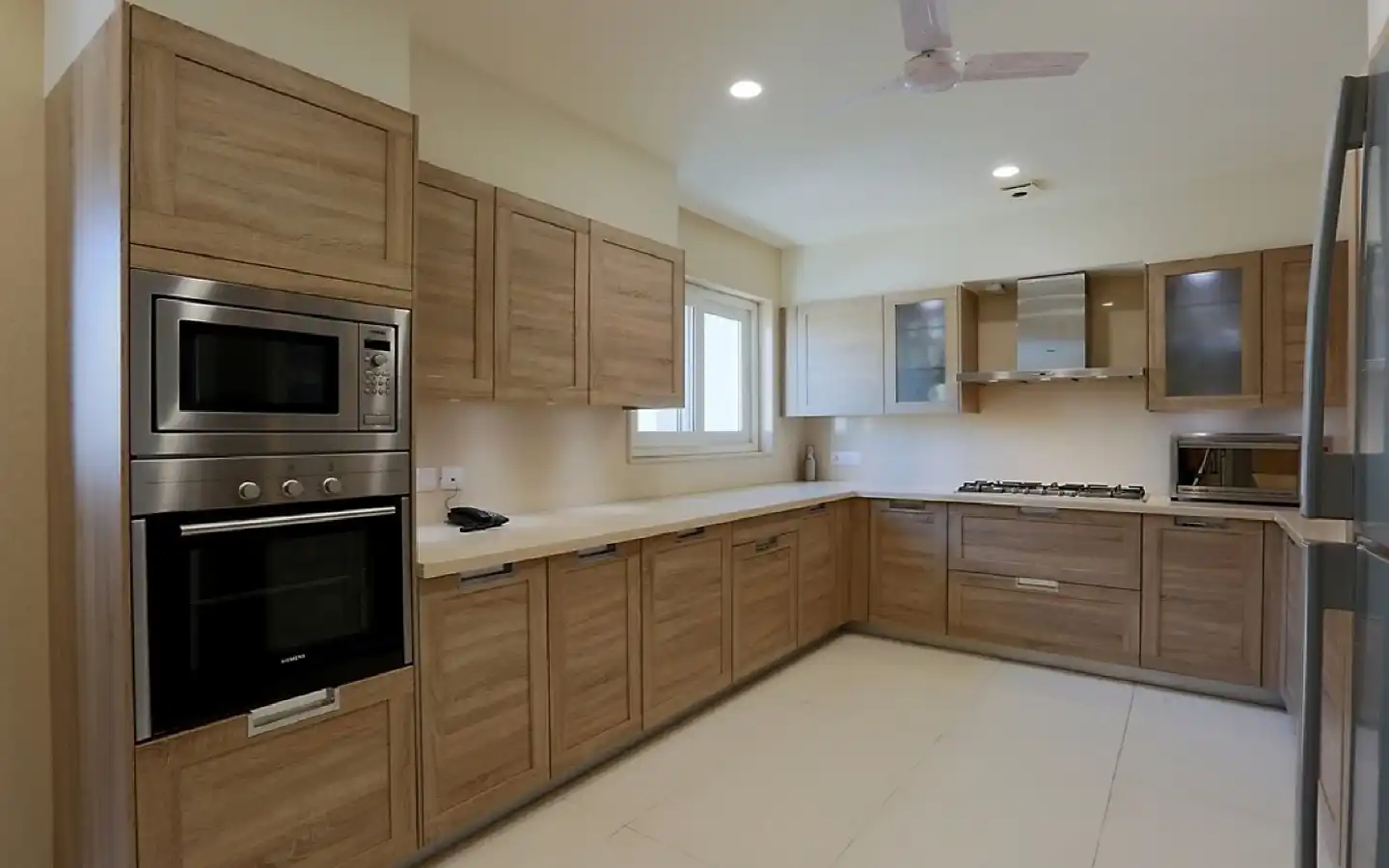 Modern L-shaped modular kitchen featuring light wood grain cabinets, integrated stainless steel appliances, a white countertop, and recessed ceiling lighting