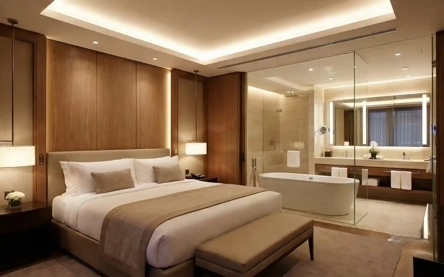 Modern luxury hotel suite interior featuring a king-size bed, warm wood paneling, and an open-concept glass-walled bathroom with a soaking tub