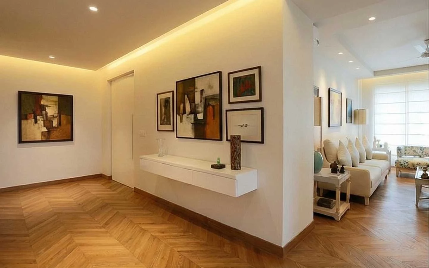 Modern minimalist interior featuring a white hallway with a floating console, abstract framed art, cove lighting, and light oak chevron pattern wood flooring leading into a bright living room
