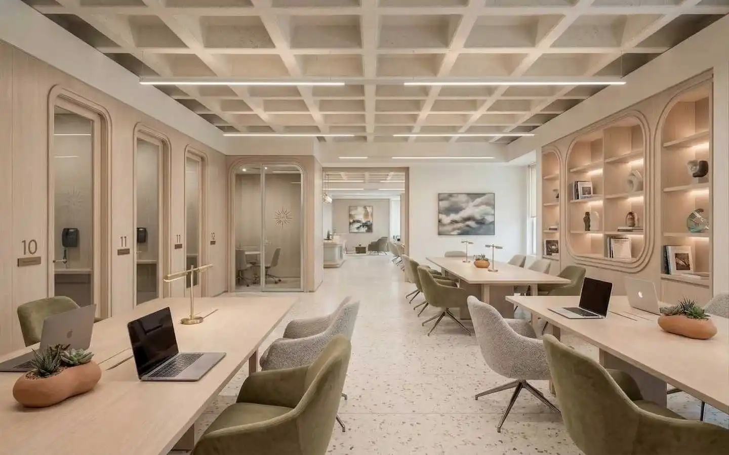 Modern office co-working space interior with long shared desks, ergonomic chairs, individual private offices numbered 10–12, laptops on desks, terrazzo flooring, and decorative shelving along the wall