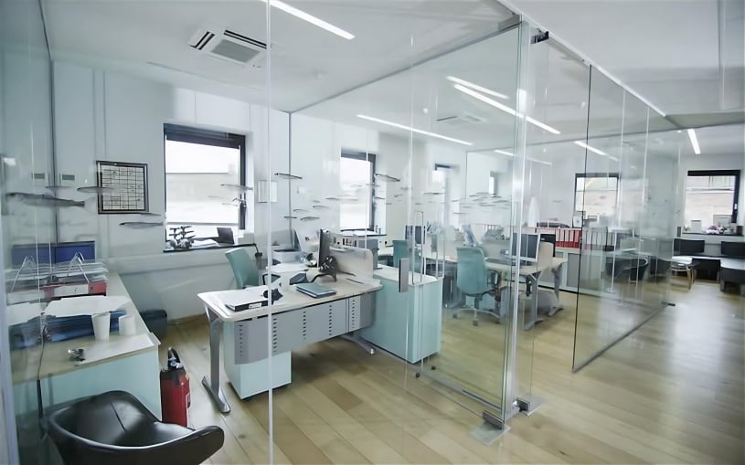 Bright open-plan office interior featuring frameless glass partitions, light wood flooring, ergonomic workstations with teal chairs, and ample natural light from large windows