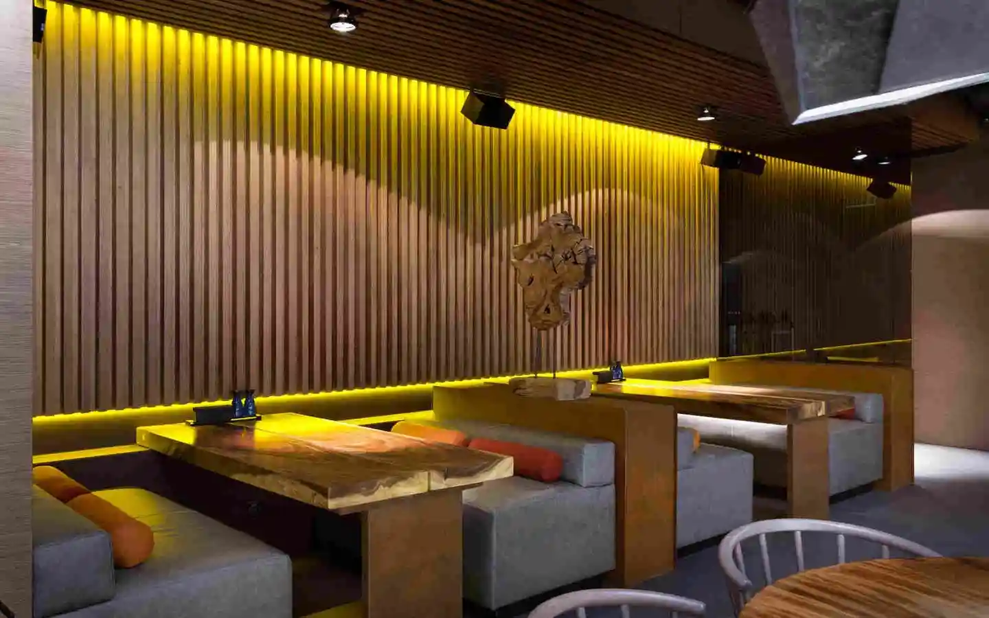 Modern restaurant interior featuring wooden fluted wall paneling with yellow cove lighting, live-edge timber tables, and plush grey booth seating
