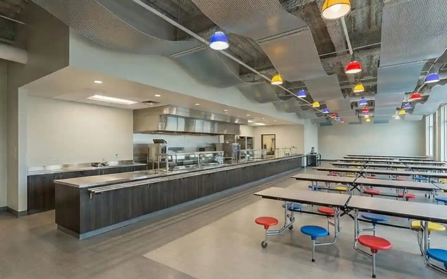 Spacious school cafeteria featuring an industrial kitchen, long dining tables with colorful stools, and a decorative wavy ceiling with multi-colored pendant lights