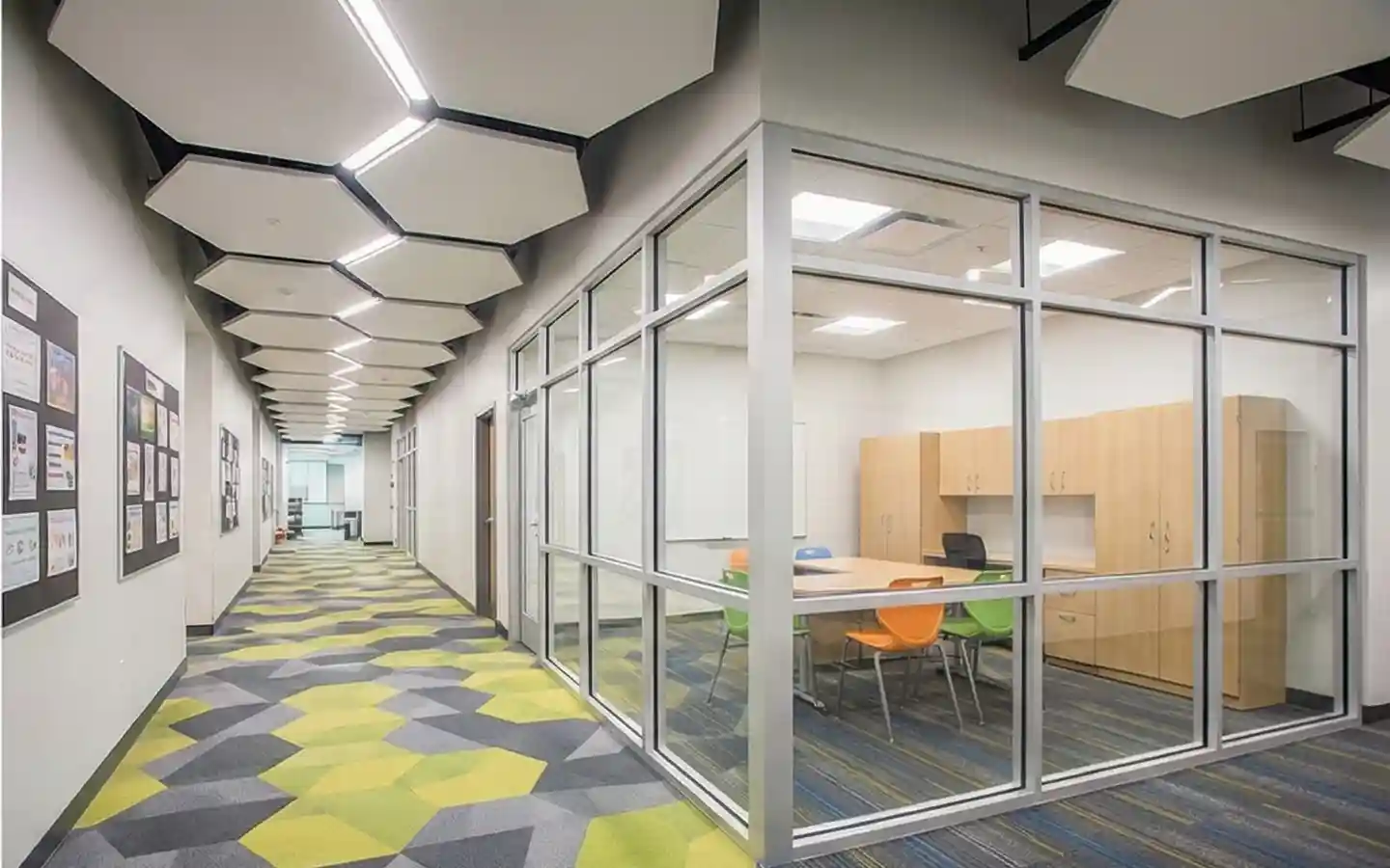 Modern school corridor featuring a geometric hexagonal ceiling, patterned carpet, and a glass-enclosed meeting room with colorful chairs
