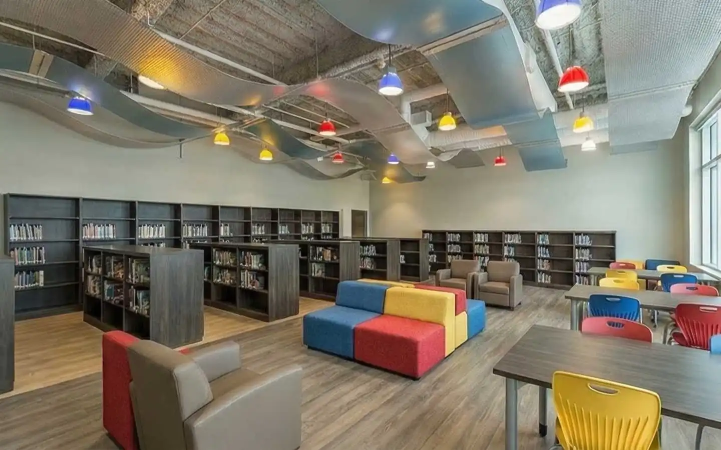 Modern school library interior featuring colorful modular seating, dark wood bookshelves, vibrant pendant lighting, and a creative wave-shaped acoustic ceiling