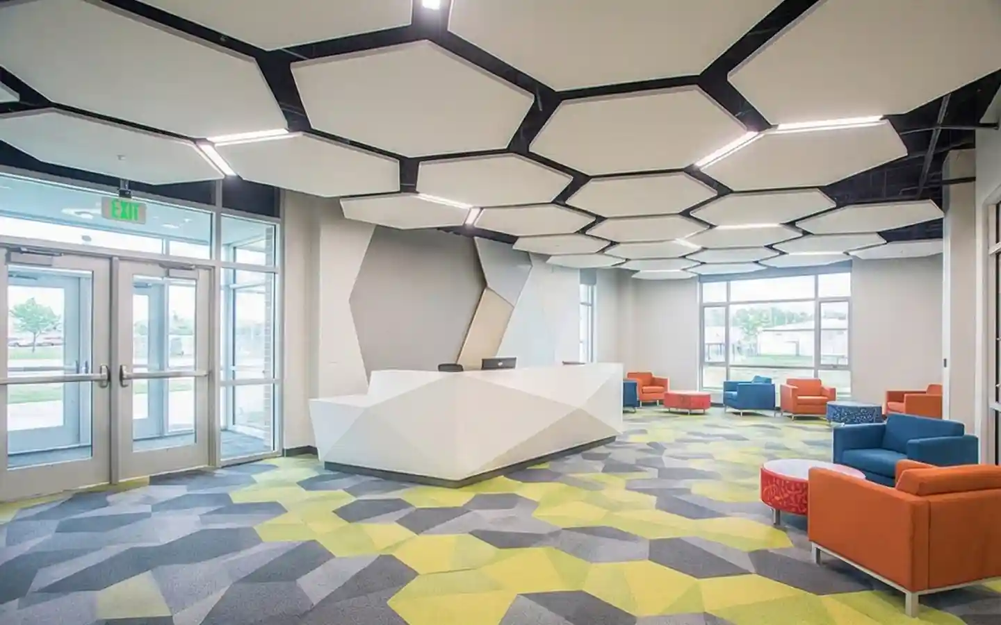 Modern school lobby interior featuring a geometric white reception desk, hexagonal acoustic ceiling panels with integrated lighting, and a colorful patterned carpet with lounge seating