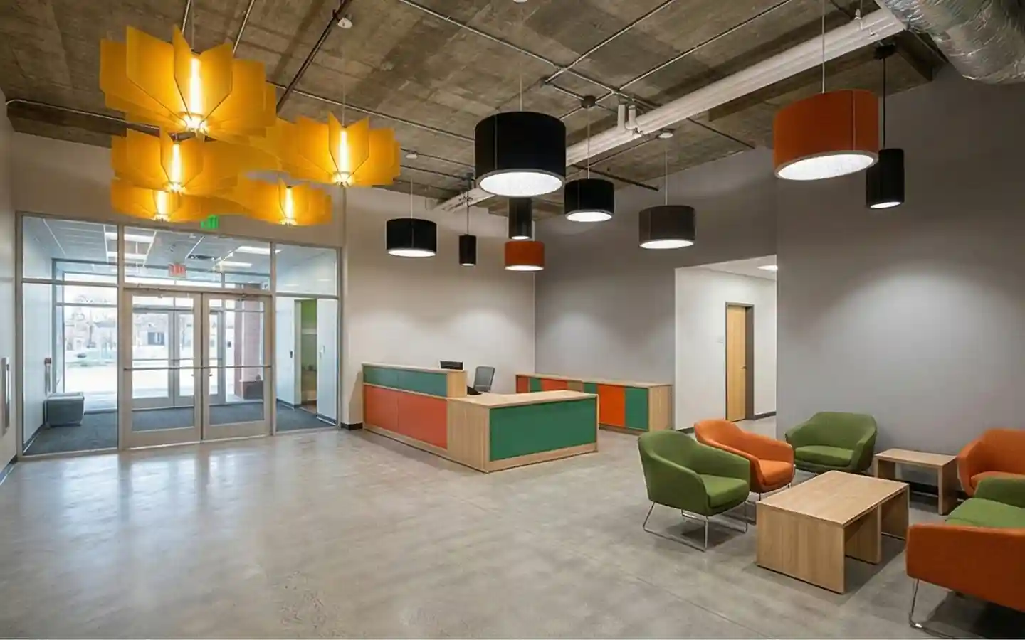 Modern school reception lobby featuring industrial concrete ceilings, colorful pendant lighting, and a contemporary waiting area with green and orange seating