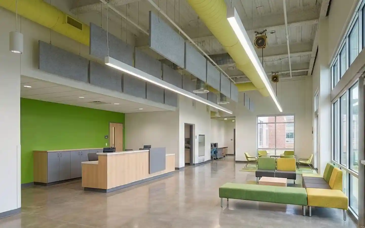 Modern school reception area with high industrial ceilings, yellow exposed ductwork, hanging acoustic baffles, and a lime green accent wall in a spacious lobby