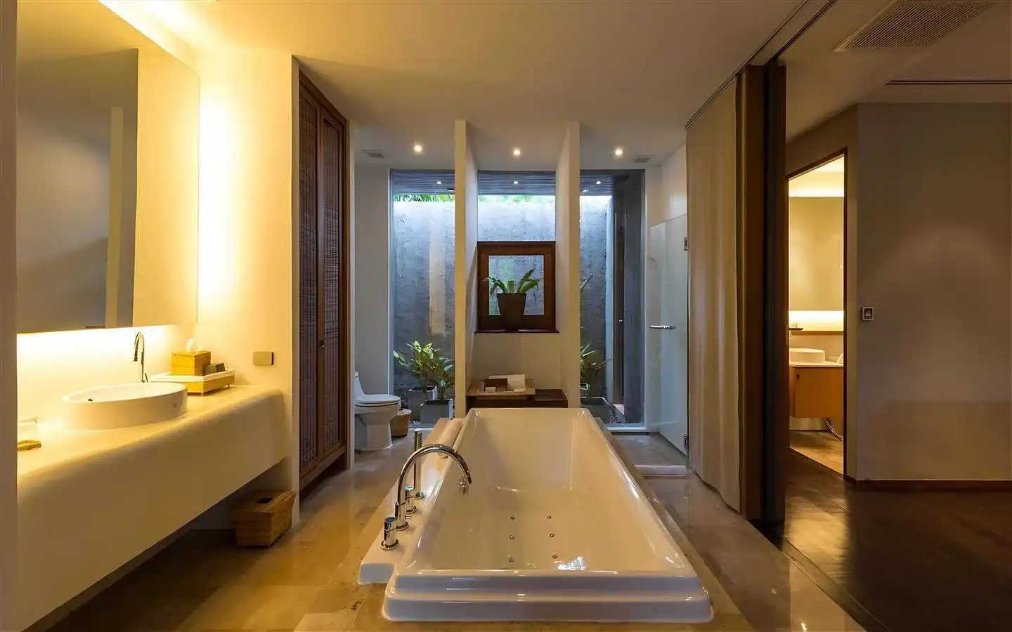 Modern spa-like bathroom interior design featuring a central freestanding jetted bathtub, a floating white vanity with warm ambient lighting, and a serene background with indoor tropical plants