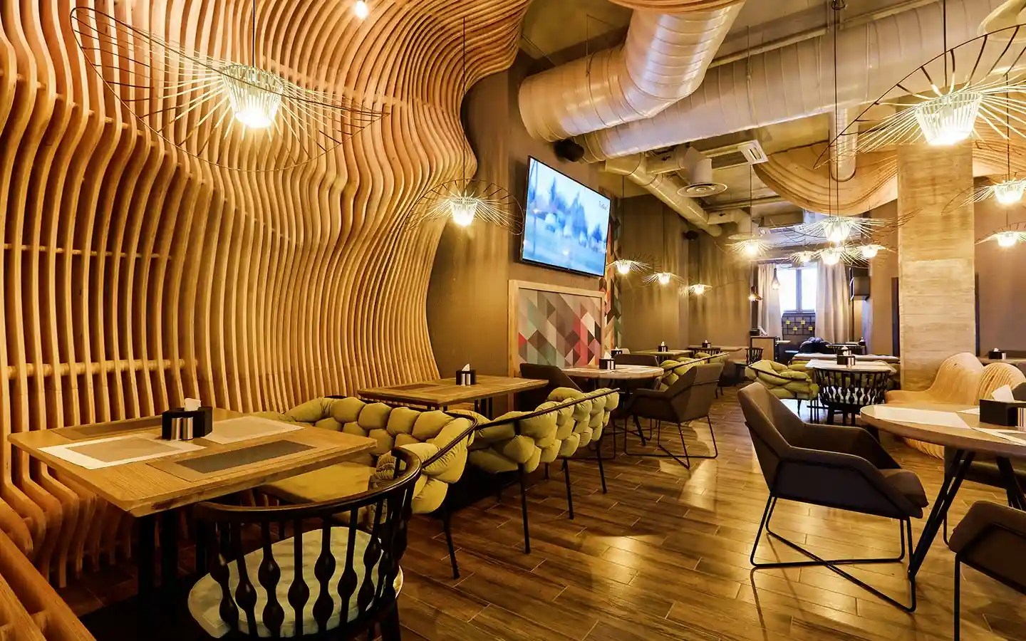 Modern restaurant interior featuring a parametric curved wood slat wall, exposed industrial ceiling ducts, and unique wire cage pendant lights over green tufted seating