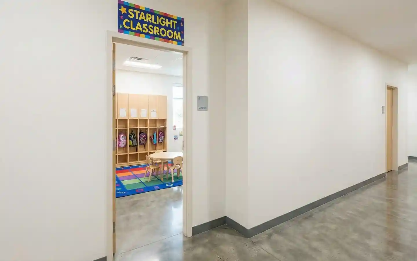 View into a preschool classroom titled Starlight Classroom featuring wooden cubby storage for backpacks, a colorful alphabet rug, and polished concrete hallway floors