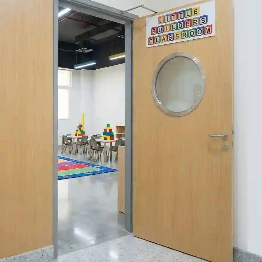 Entrance to a preschool 'Little Builders' classroom featuring a light wood door with a circular porthole window and a bright activity area inside
